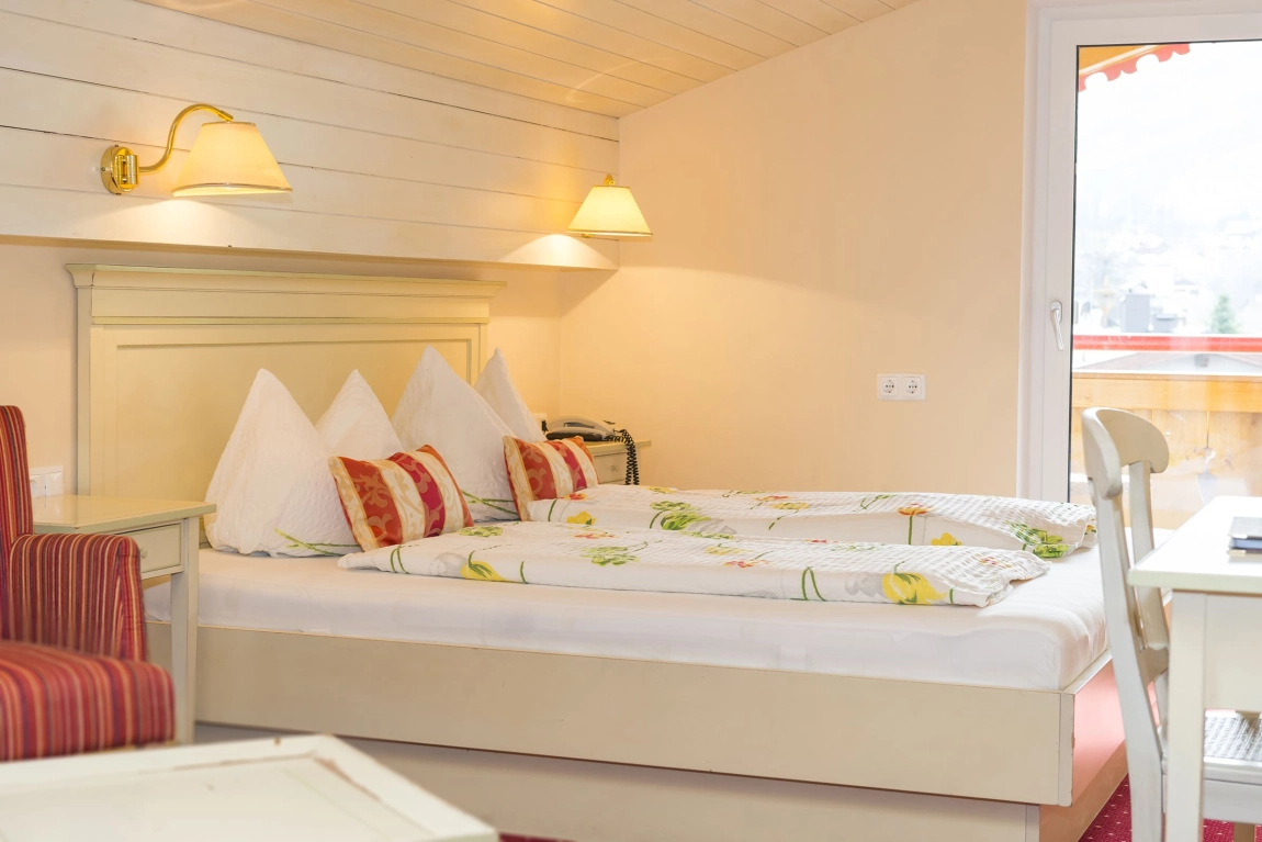 Gemütliches Doppelbett mit Blumenbettwäsche und Zierkissen, seitlich Schreibtisch und Zugang zum Balkon mit Holzbrüstung.
